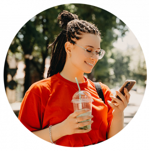 A woman with dark hair, glasses, and a red shirt holding a drink in one hand and her mobile phone in the other hand